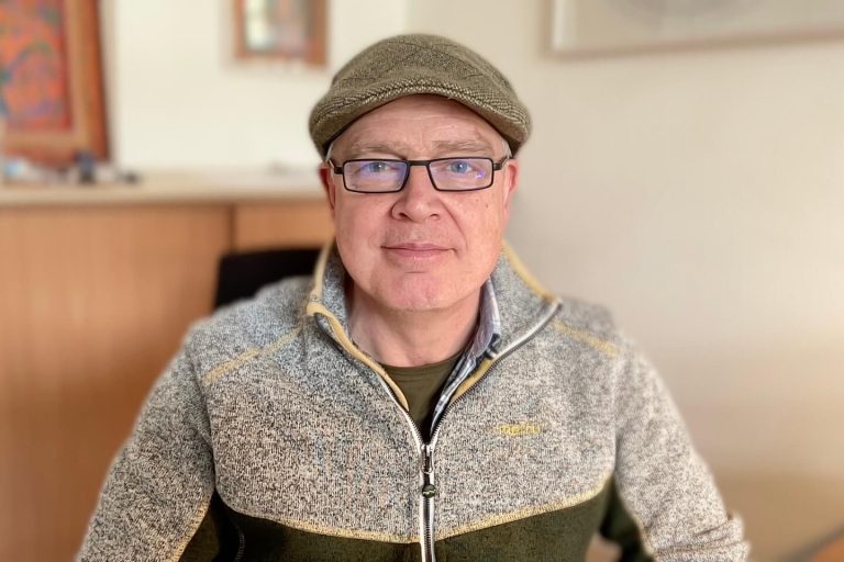 Mann mit Brille und Filzhut in einem hellen Raum, trägt einen grauen Pullover.