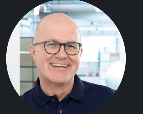 Lächelnder Mann mit Brille, kurz geschorenes Haar, in einem modernen Büro.